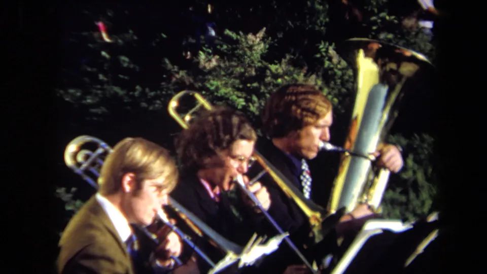 Three young men continue playing brass instruments in a 1971 Sacramento outdoor performance, framed with warm 8mm color and foliage.
