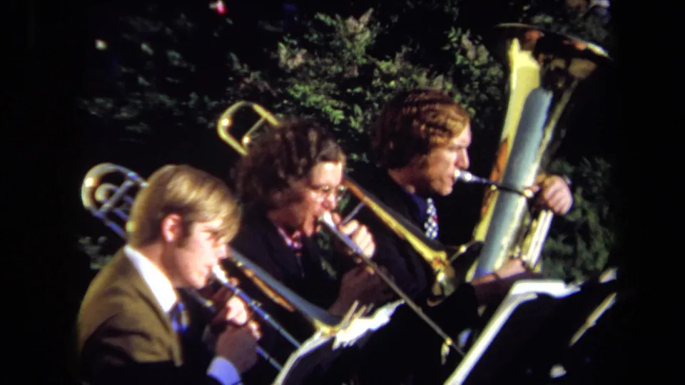 A profile view from the 1971 Sacramento home movie shows three suited brass players performing beside music stands outdoors.