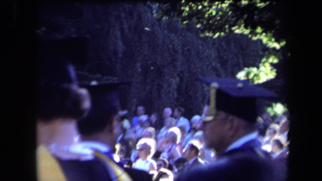 1971 Sacramento home-movie frame showing graduates in caps and gowns near a seated commencement audience under leafy trees.