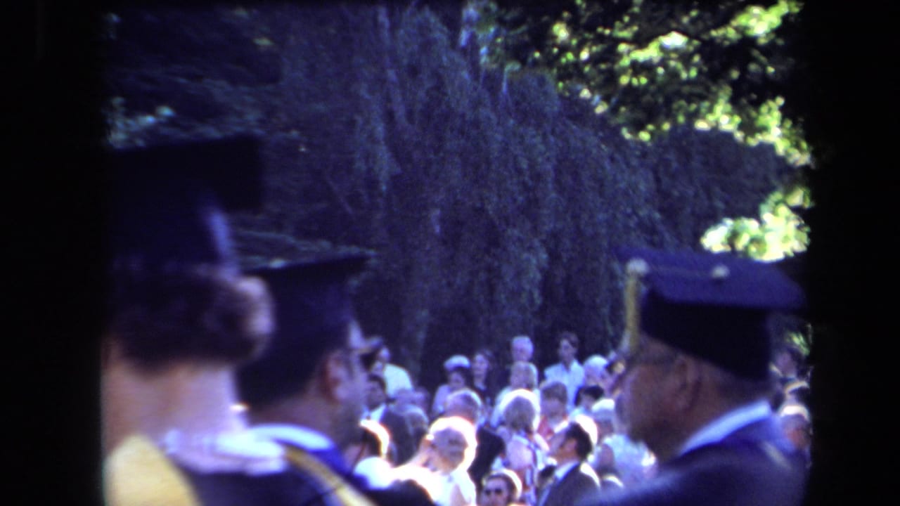 Home-movie view of graduates in academic dress close to the camera, with a Sacramento commencement audience behind them.