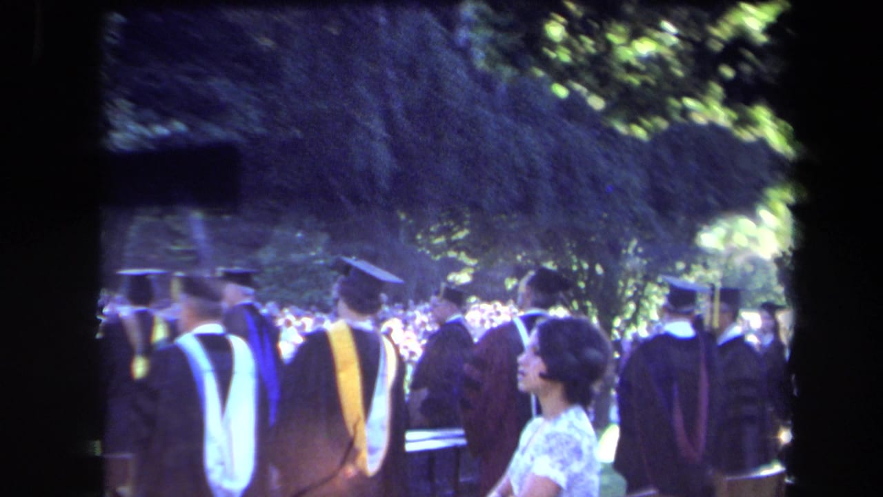 1971 Sacramento commencement frame showing graduates walking past a white-dressed attendee and a crowd under trees.
