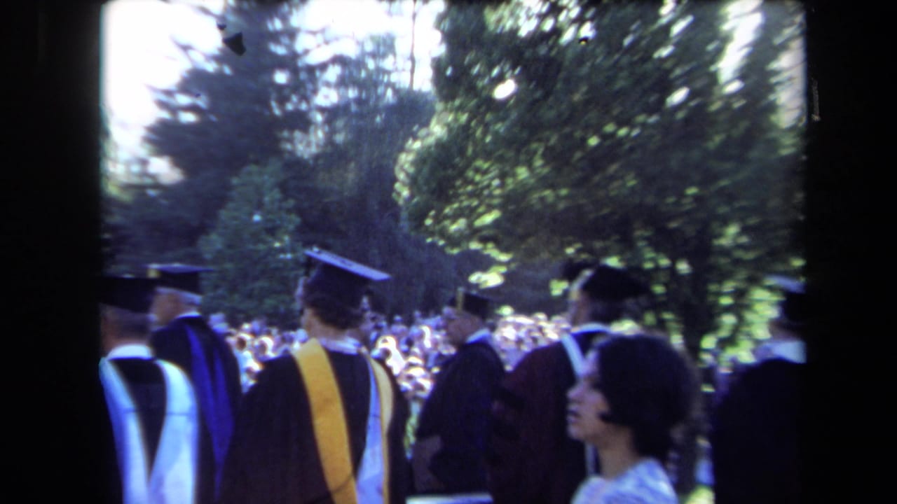 1971 Sacramento home-movie frame showing graduates in mortarboards and gowns near spectators under trees.