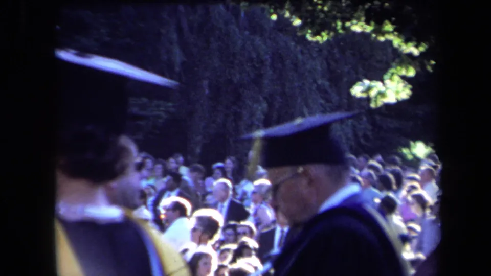 1971 outdoor graduation footage showing figures in academic dress moving past a sunlit seated audience.