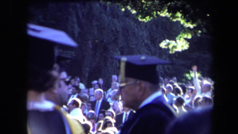 Sacramento graduation home-movie frame with caps and gowns in the foreground and spectators seated outdoors.