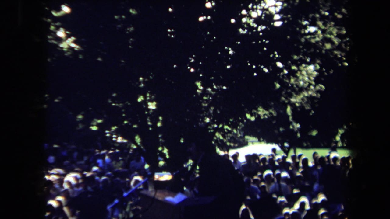 Nighttime 8mm frame from Sacramento showing an outdoor audience beneath trees with blue light and a small illuminated area near the stage.