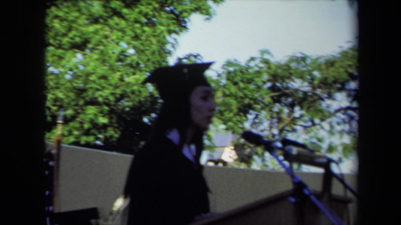 Graduate in cap and gown stands at an outdoor podium near a microphone.