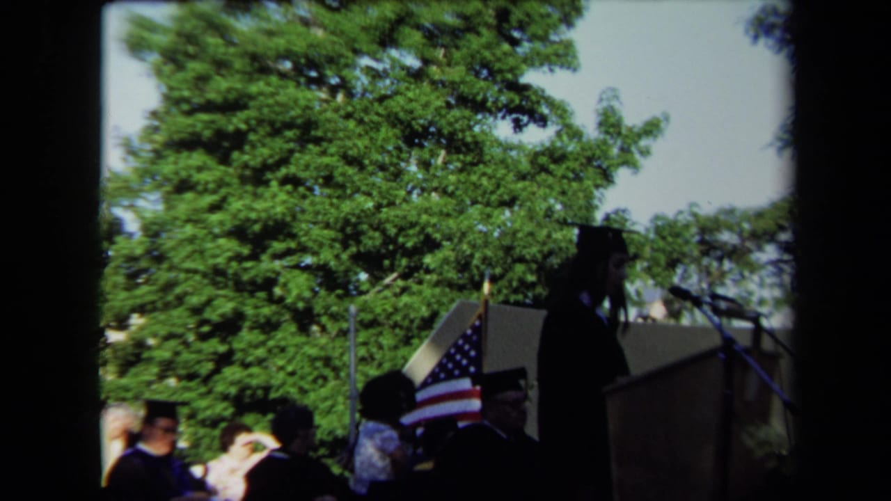 Outdoor graduation stage in Sacramento, 1971, with a speaker, American flag, and seated attendees visible.