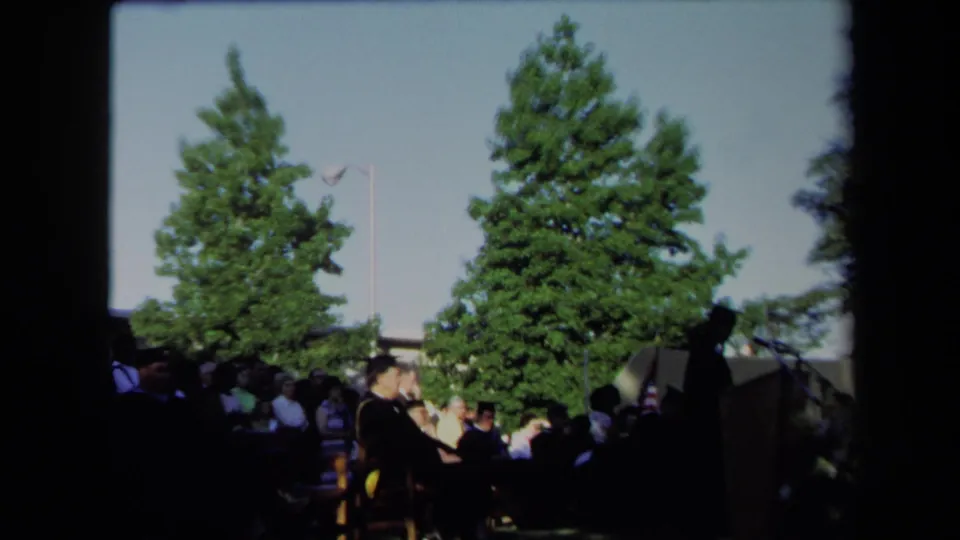 Wide 8mm view of a Sacramento graduation ceremony in 1971 with attendees, trees, and a flag by the stage.