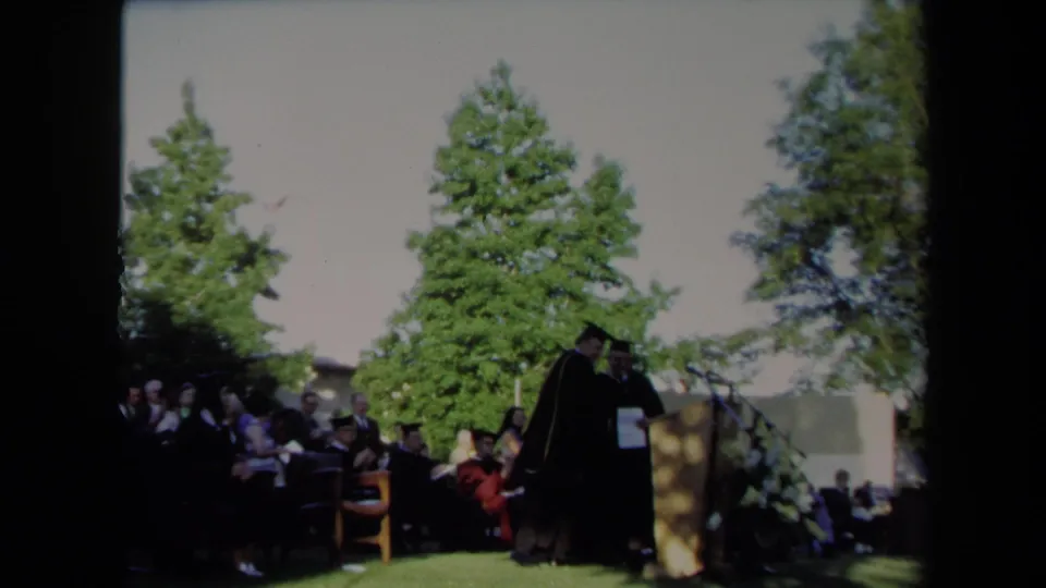Graduation participants in robes move near the podium as seated guests watch outdoors in Sacramento.