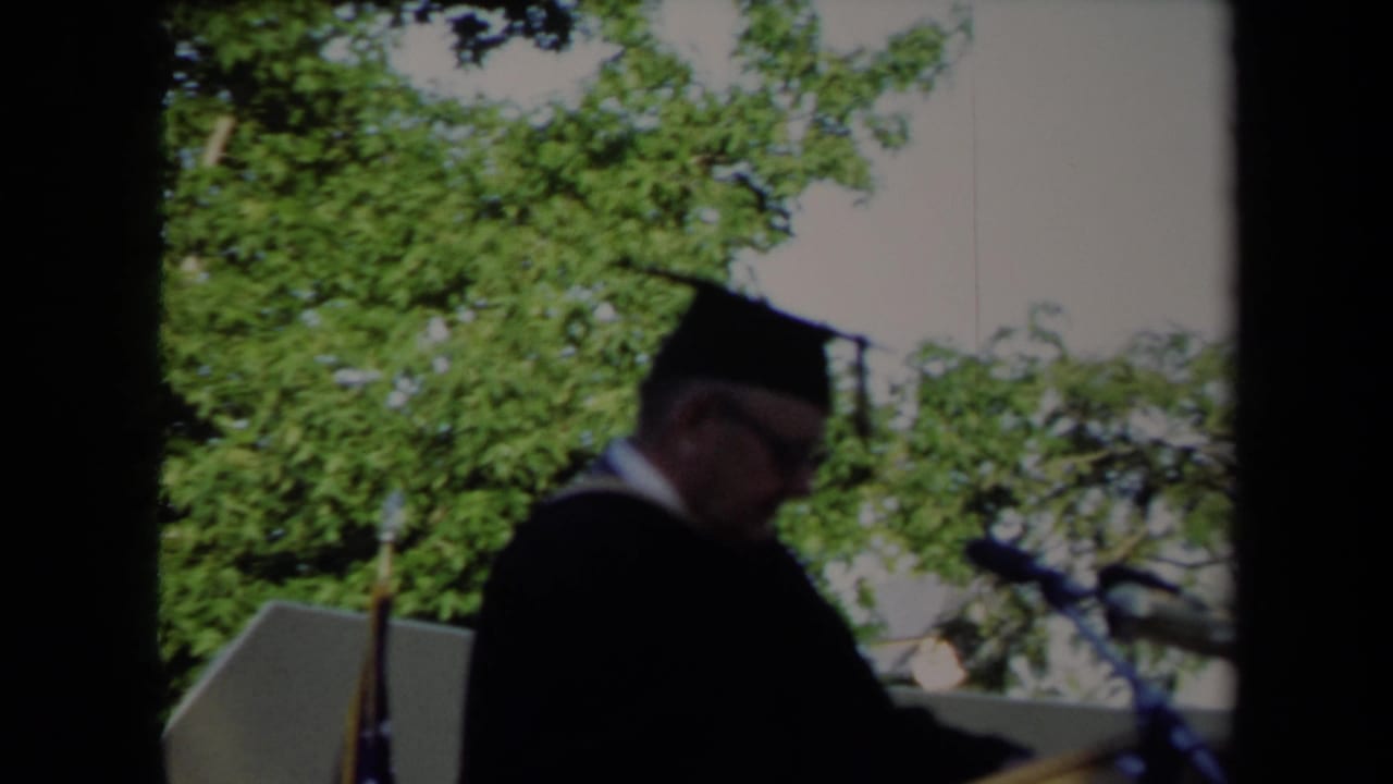 Side view of a man in academic regalia at an outdoor Sacramento graduation podium in 1971, with trees and bright sky behind him.