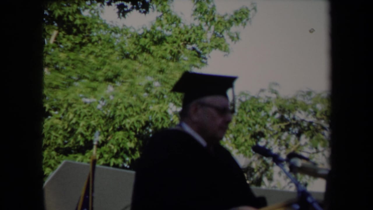 Profile view of the 1971 Sacramento graduation speaker in cap and gown, positioned at a podium under daylight.