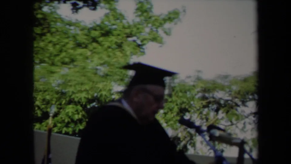 Soft 8mm frame of a 1971 Sacramento commencement speaker at an outdoor podium, partly blurred by camera movement.