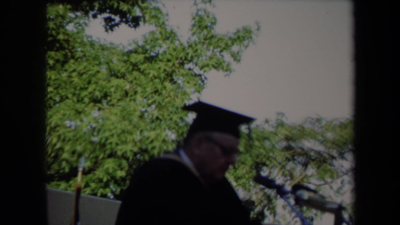 The 1971 Sacramento graduation speaker remains near the microphones, framed by leafy trees and bright daylight.