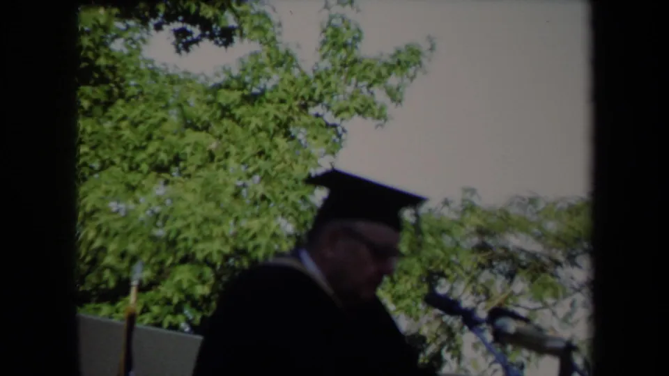 The 1971 Sacramento graduation speaker remains near the microphones, framed by leafy trees and bright daylight.