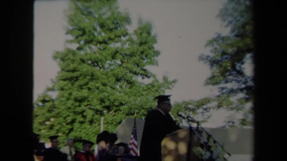 A wider 8mm view shows the Sacramento graduation speaker at a podium with seated graduates and an American flag visible nearby.
