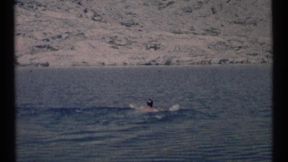 A distant man swims near the right side of a calm Montana lake, framed by 8mm film borders and a rocky hillside.