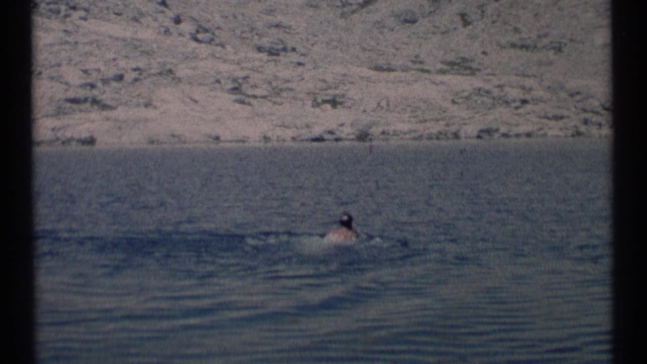 A distant adult swimmer moves through a lake in Scapegoat Wilderness, Montana, with a rocky hillside in the background.