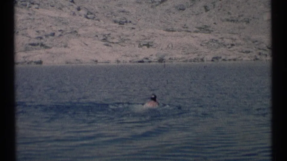 A distant adult swimmer moves through a lake in Scapegoat Wilderness, Montana, with a rocky hillside in the background.