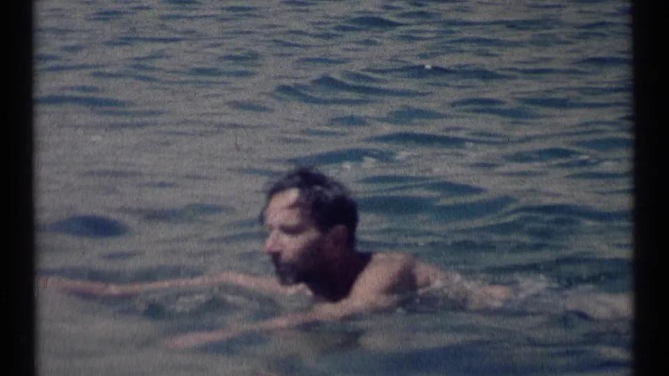 The dark-haired man continues swimming across the lake, partly low in frame, in soft 8mm color.