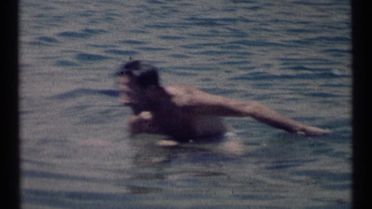 A man swims through calm lake water in 1973 Scapegoat Wilderness home-movie footage, framed in soft, grainy 8mm texture.