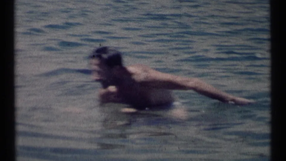 A man swims through calm lake water in 1973 Scapegoat Wilderness home-movie footage, framed in soft, grainy 8mm texture.