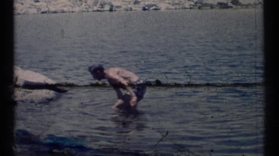 A man wades near a rocky lakeshore in 1973 Scapegoat Wilderness footage, with muted color and handheld home-movie framing.
