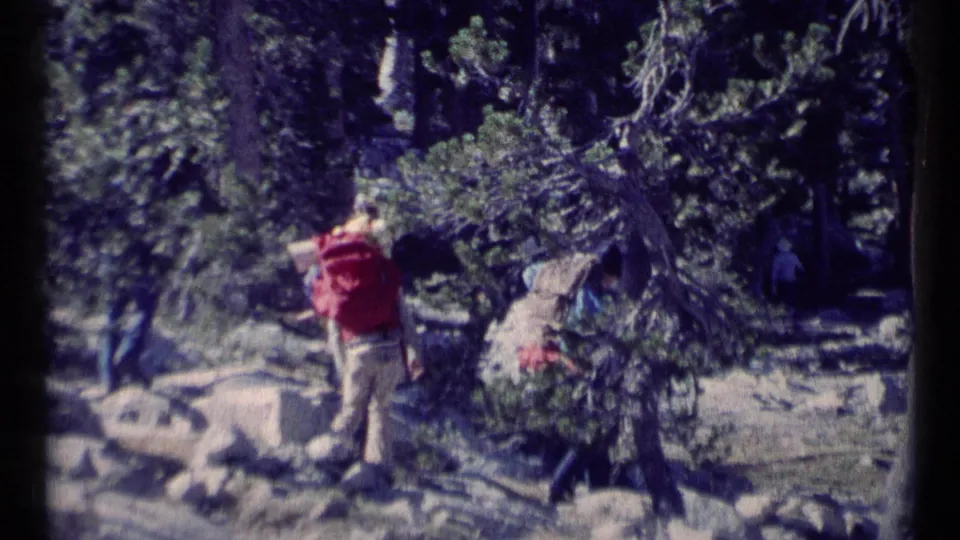 Hikers with backpacks move through a wooded rocky trail in 1973 Scapegoat Wilderness home-movie footage.