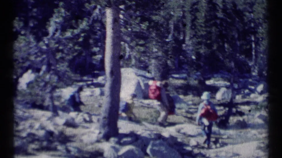 1973 8mm Montana wilderness footage shows backpacked hikers moving through trees and rocks on uneven ground.