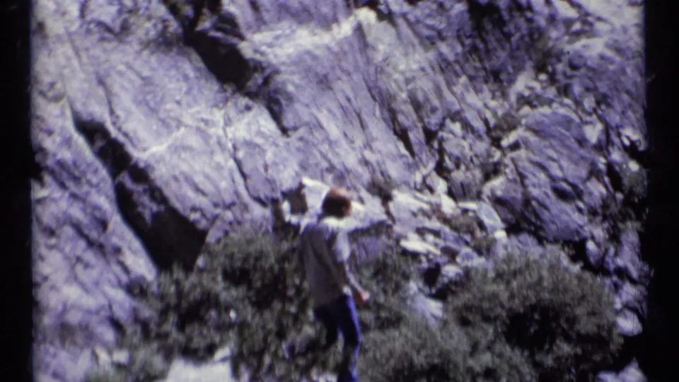 In 1973 Montana wilderness footage, a hiker in a hat stands near scrub vegetation below a large rock face.