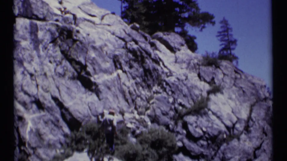 A distant hiker appears below a broad gray rock formation in Scapegoat Wilderness, Montana, on 8mm film.