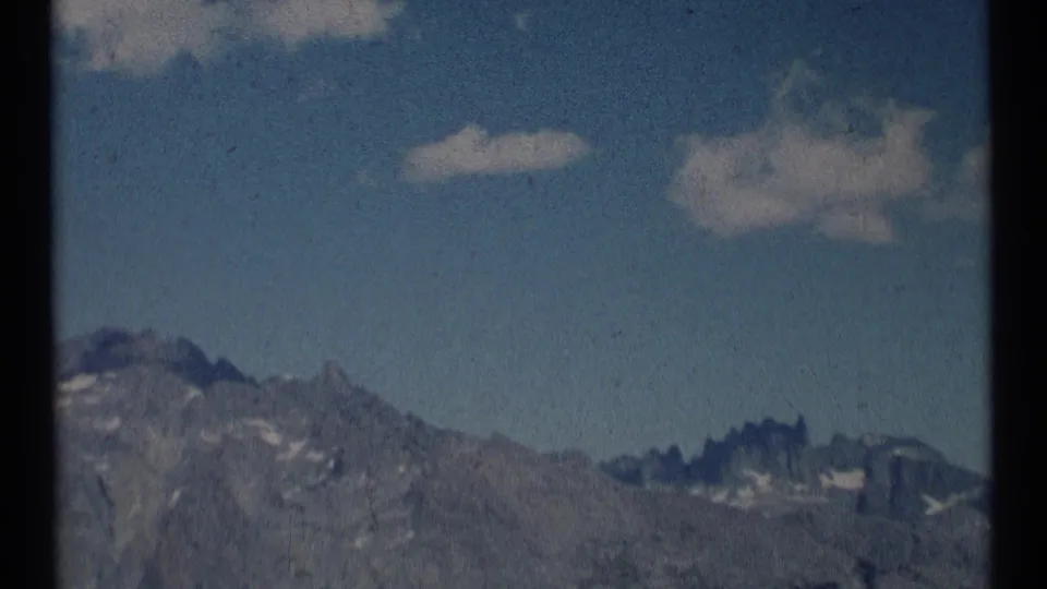 1973 home-movie frame from Scapegoat Wilderness showing jagged mountain peaks, snow patches, and open blue sky.