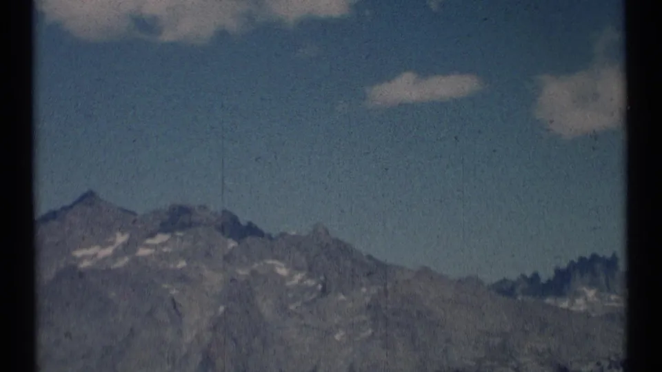 8mm 1973 Montana wilderness frame showing gray mountain slopes, scattered snow, and pale clouds in blue sky.