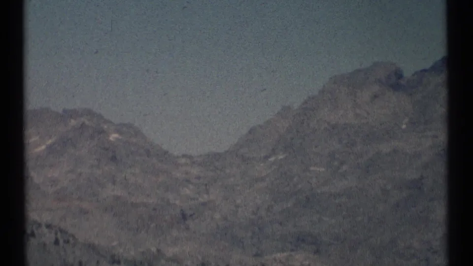 The 8mm view tightens on rugged Scapegoat Wilderness mountains, showing a broad rocky peak and subdued 1970s film color.