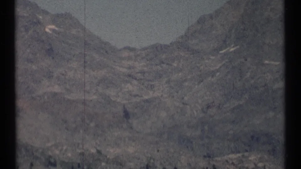 A closer 1973 8mm view of Montana wilderness terrain shows rocky slopes, small snow remnants, and soft home-movie focus.