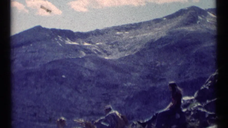 1973 8mm Montana wilderness view with snow-streaked peaks and a person seated on rocks looking toward the mountains.