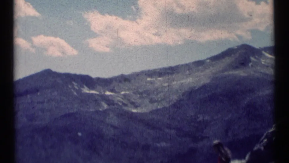 1973 8mm view of Scapegoat Wilderness mountains beneath scattered clouds, with part of a seated person visible near the rocks.