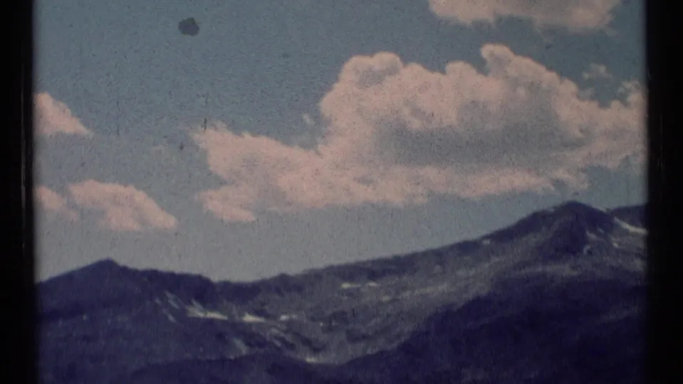 1973 8mm Montana mountain vista showing ridges, lingering snow patches, and bright clouds over the wilderness landscape.