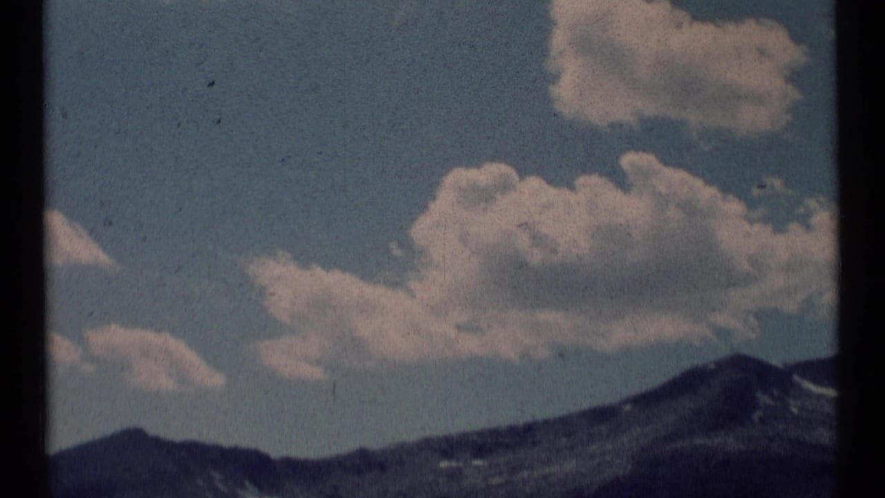 1973 8mm frame from Scapegoat Wilderness, Montana, showing cumulus clouds over dark mountain peaks under a blue sky.