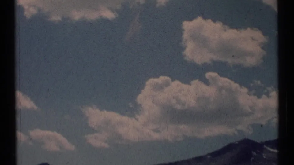 1973 8mm frame showing white clouds above a sloped mountain ridge in Scapegoat Wilderness, Montana.