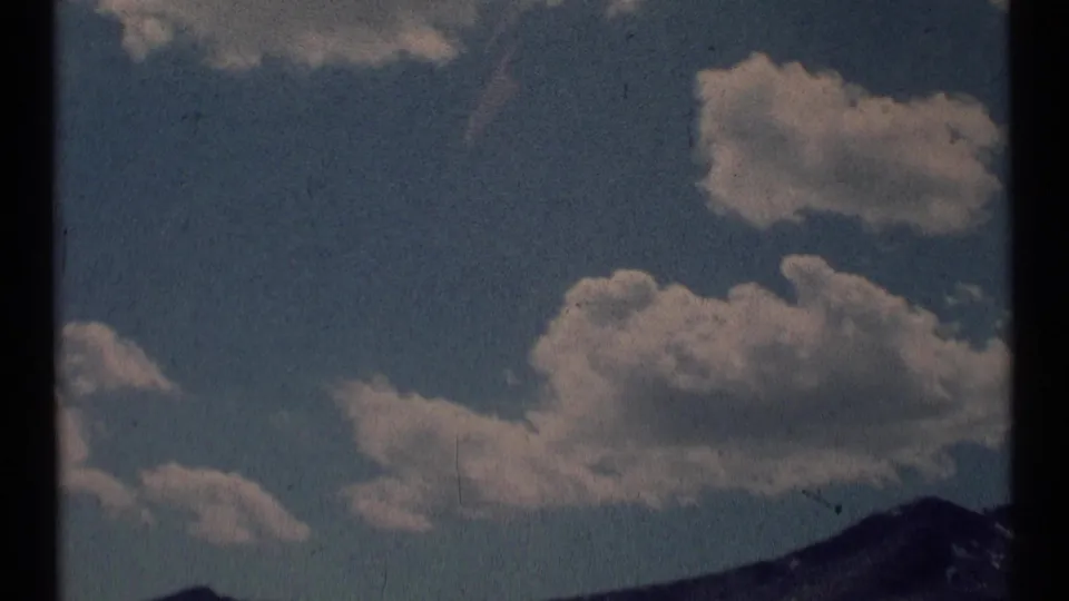 1973 8mm Montana mountain-sky frame with clustered cumulus clouds above a dark ridge at the lower edge.