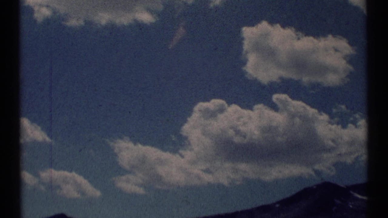 1973 8mm home-movie frame from Scapegoat Wilderness, Montana, showing white clouds over a dark mountain horizon.