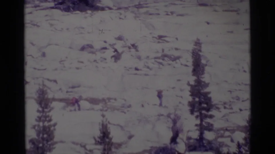 Wide home-movie frame of hikers, one wearing red, crossing rocky Minaret Wilderness terrain under hazy exposure.