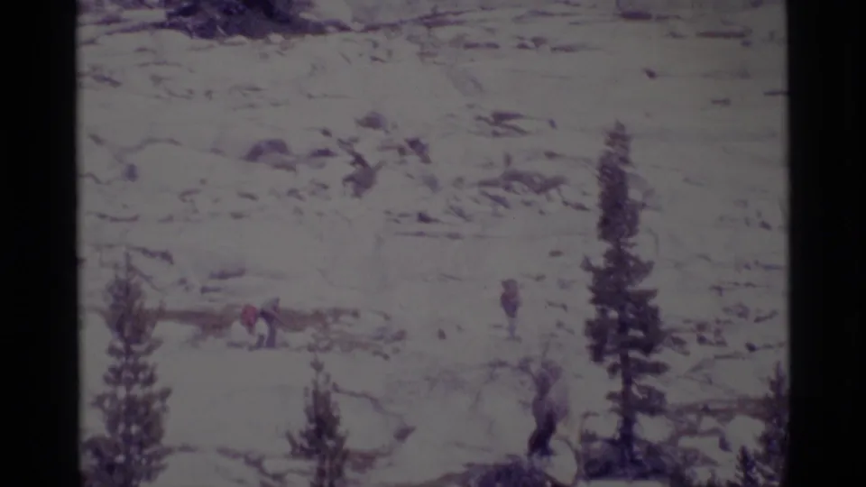 1975 8mm view of small hikers with backpacks moving through Minaret Wilderness terrain with rocks and scattered conifers.
