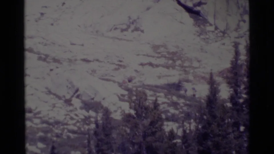 Soft 8mm frame of Minaret Wilderness terrain showing rocks, pale ground, and foreground conifers with hikers hard to distinguish.
