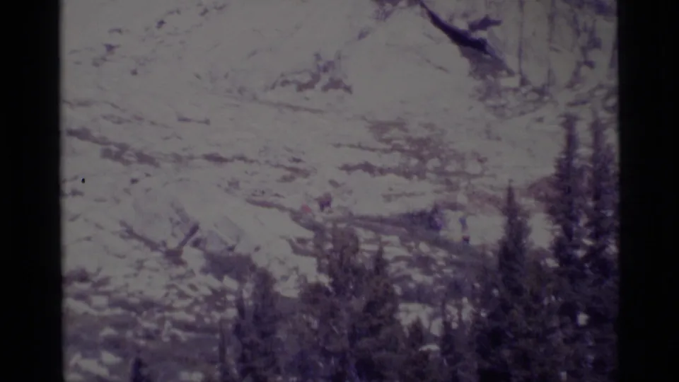 1975 home-movie frame of Minaret Wilderness terrain with scattered trees and faint distant hikers in a bright rocky landscape.