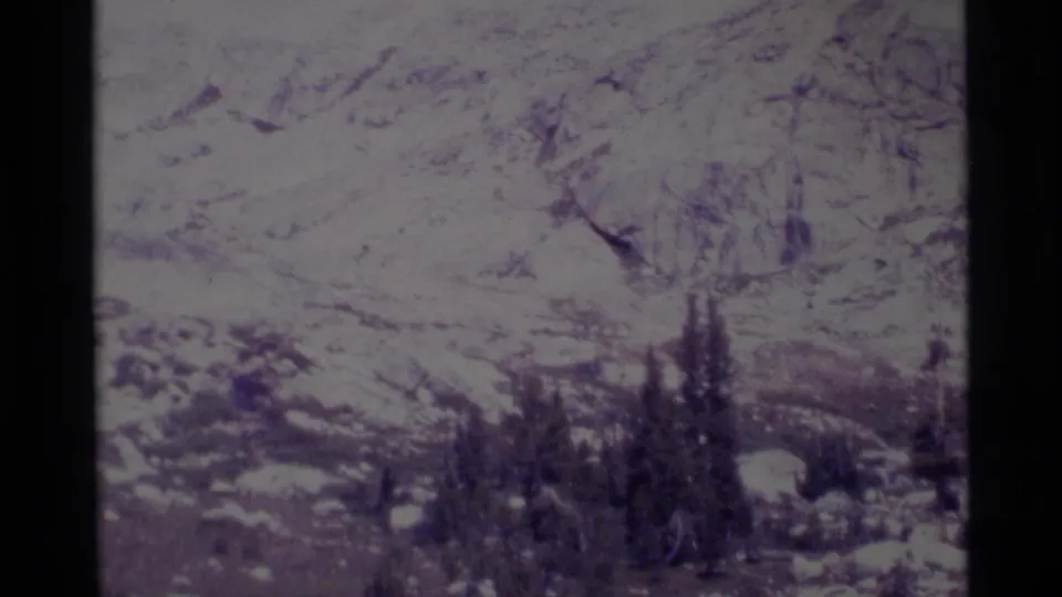 A grainy 1975 8mm frame shows snowfields, rugged slopes, and dark conifers in California's Minaret Wilderness setting.