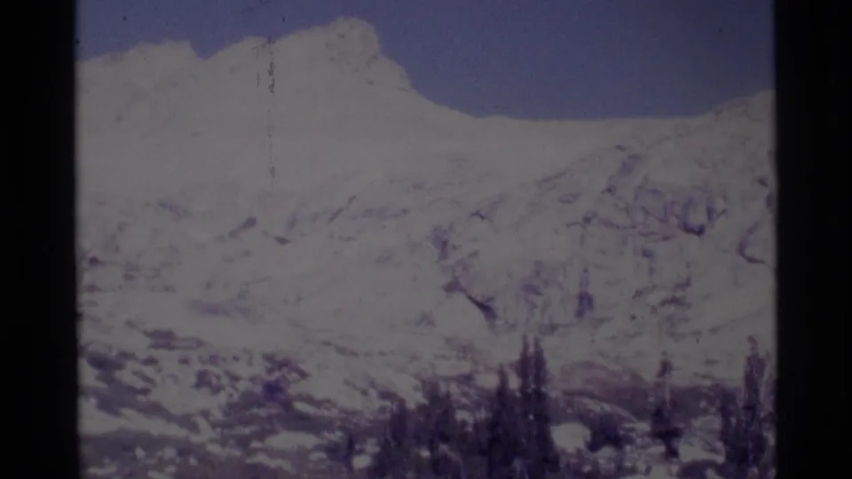 1975 8mm Minaret Wilderness view of a snowy mountain ridge, exposed rock, blue sky, and dark evergreen trees below.