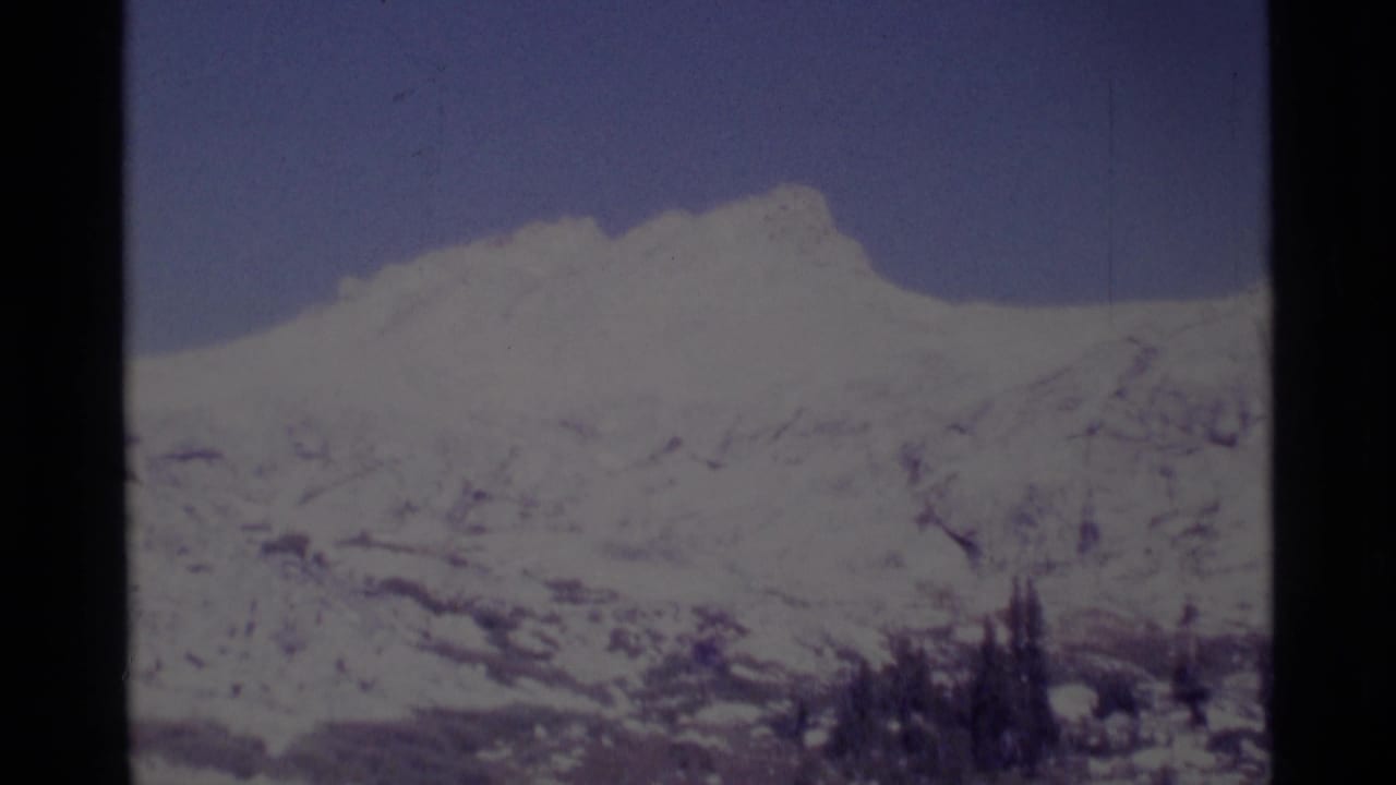 1975 8mm California wilderness frame showing a distant snow-covered mountain ridge, pale blue sky, and clustered evergreens below.