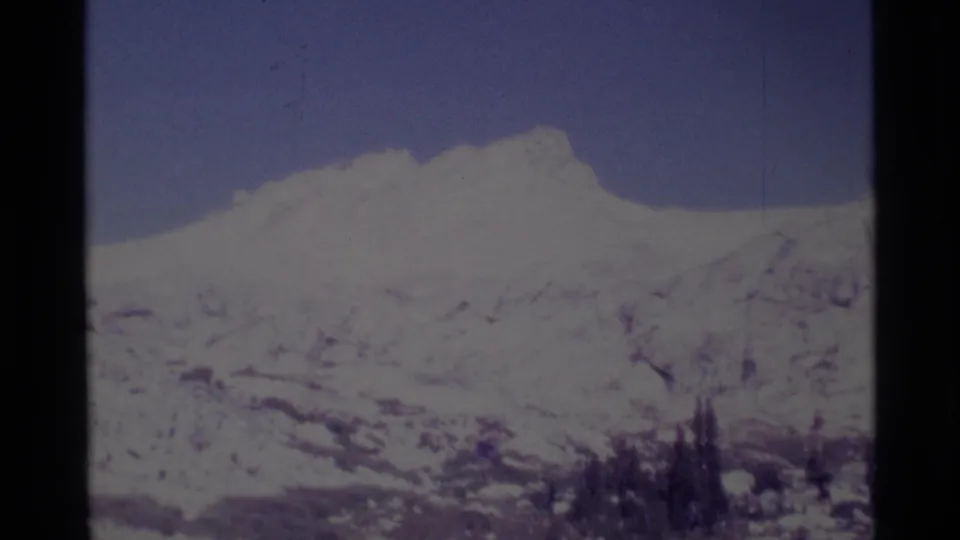 1975 8mm California wilderness frame showing a distant snow-covered mountain ridge, pale blue sky, and clustered evergreens below.