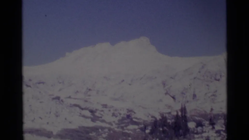 Soft 1975 8mm view of a snow-bright Minaret Wilderness ridge, muted blue sky, lower rocky slopes, and small evergreen silhouettes.